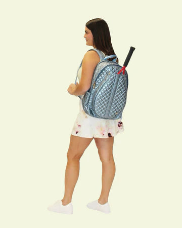 A woman in a tennis dress carrying a frosted sky blue tennis bag with multiple compartments, featuring a quilted design and a racquet attached to the top handle.