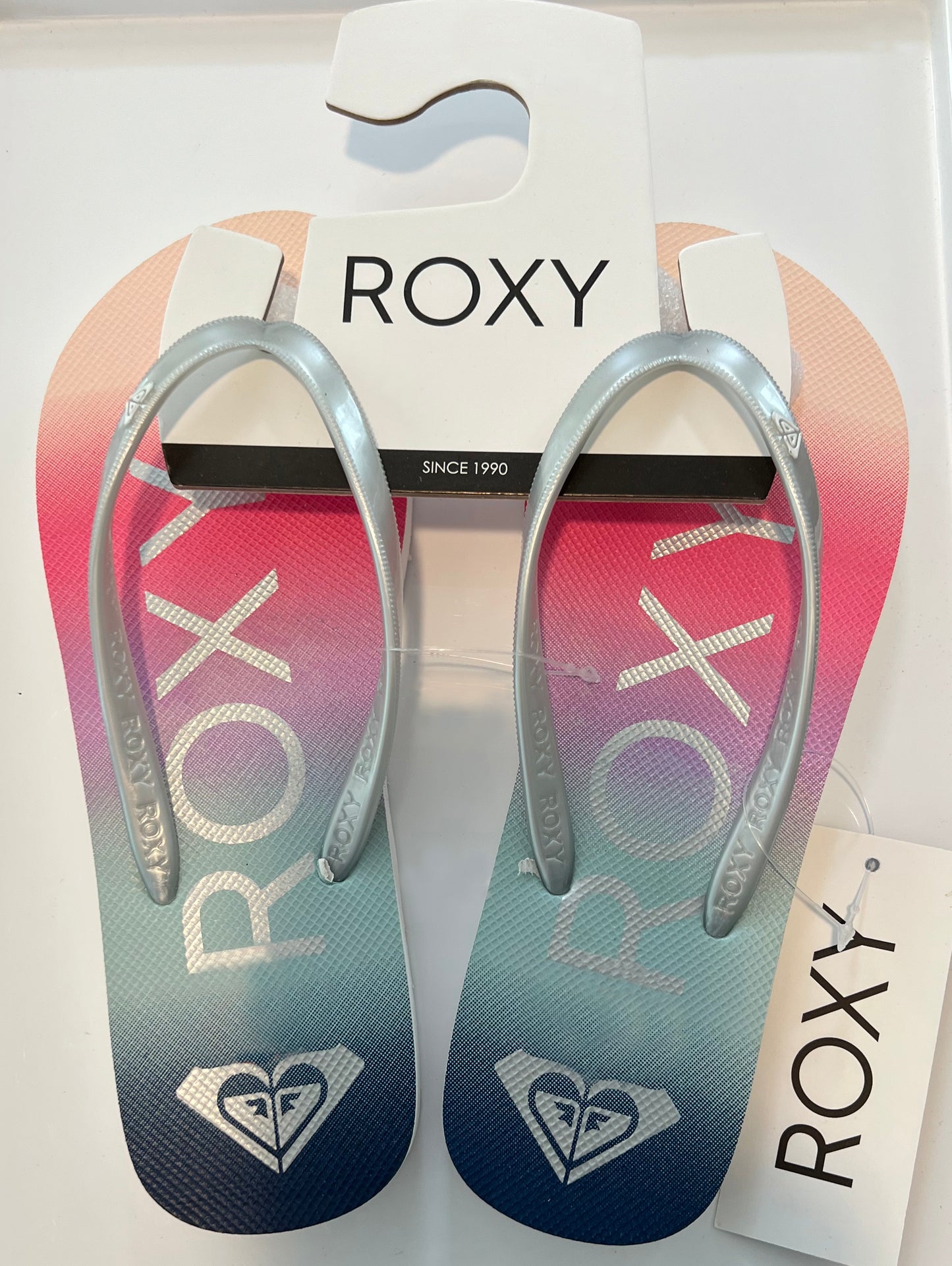 A pair of Roxy branded flip-flops with a gradient of pink to silver color, featuring the Roxy logo and a heart symbol on the straps.
