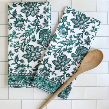 A pair of green floral patterned kitchen towels hanging on a hook with a wooden spoon beside them, displayed on a white tile background.