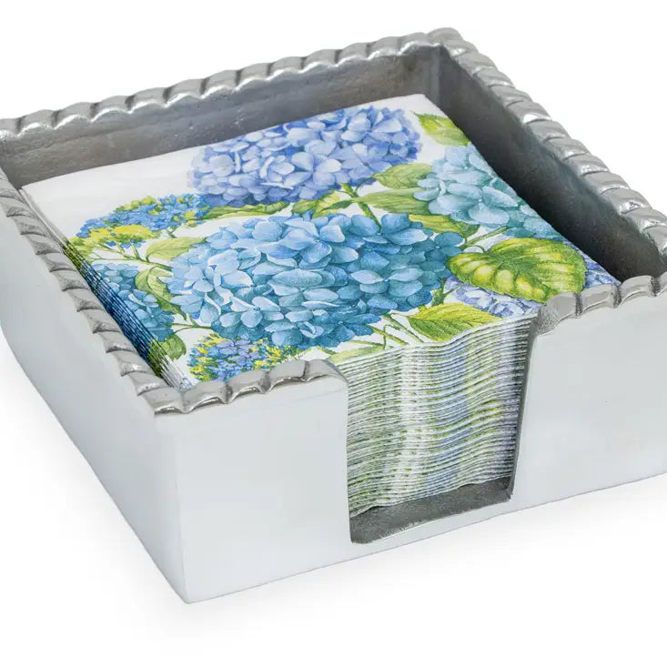 A silver metal cocktail napkin caddy with rope edge design filled with colorful cocktail napkins featuring a floral pattern.