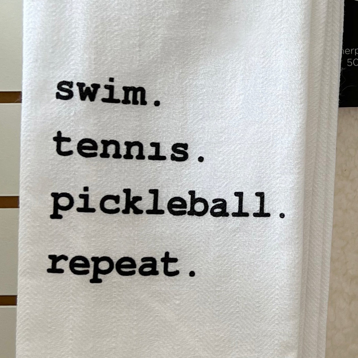 A white cotton tea towel with the words 'SWIM, TENNIS, PICKBALL, REPEAT.' printed in black lettering.
