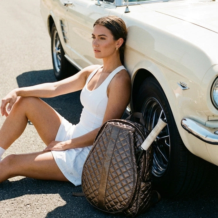 model wearing white tennis dress with rackets in cappuccino colored tennis backpack sitting leaning against car